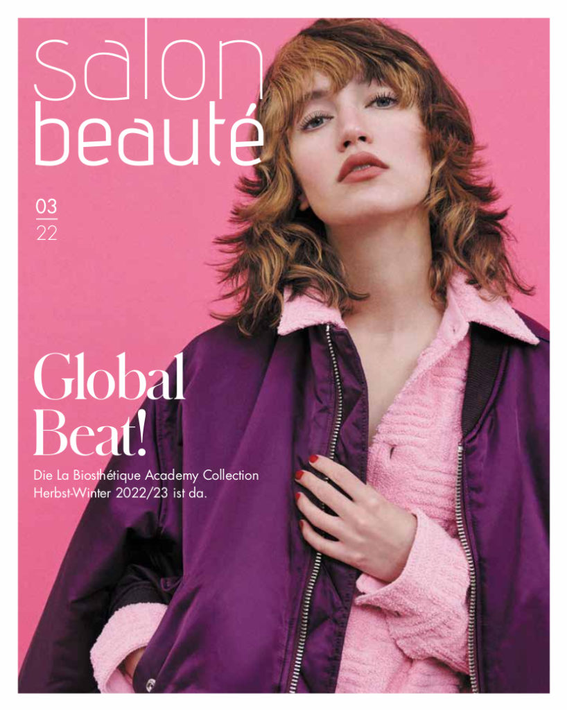 Titelseite der Zeitschrift. Eine Frau mit braunen gelockten Haar posiert und schaut von oben herab in die Kamera. Hintergrund und Strickpullover ist rosa, die Jacke violett.